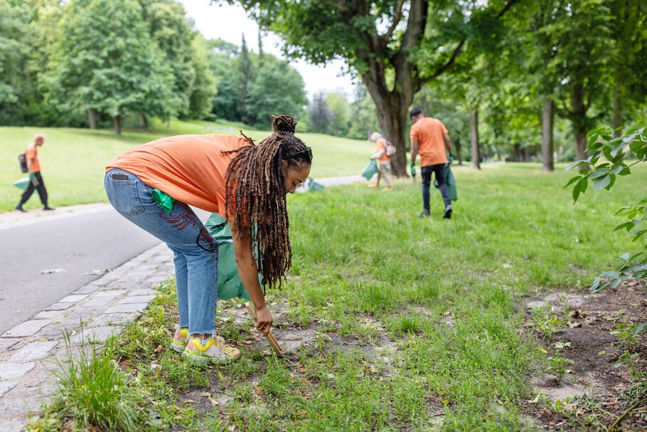 A woman wearing jeans and an orange t-shirt stands outside holding a trash bag and bends over to pick up trash with tongs. People in orange t-shirts walk around in the background.